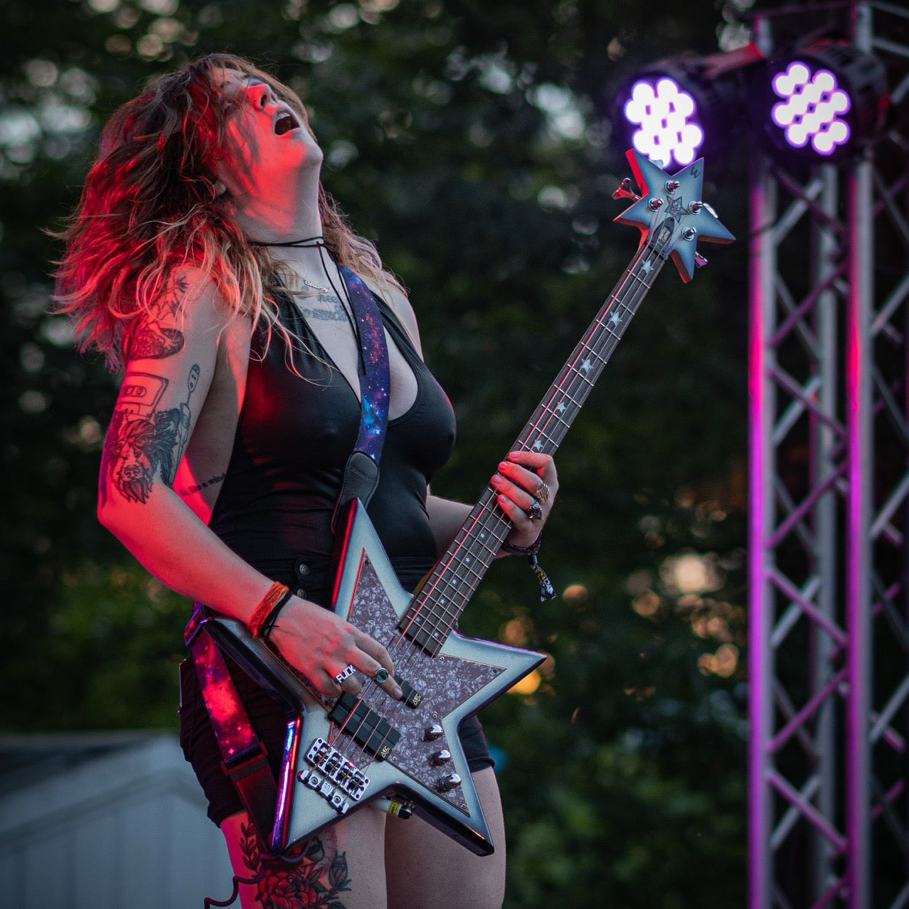 Image shows a person playing guitar on stage. They are dressed in leather with long hair and tattoos. The guitar is star shaped.